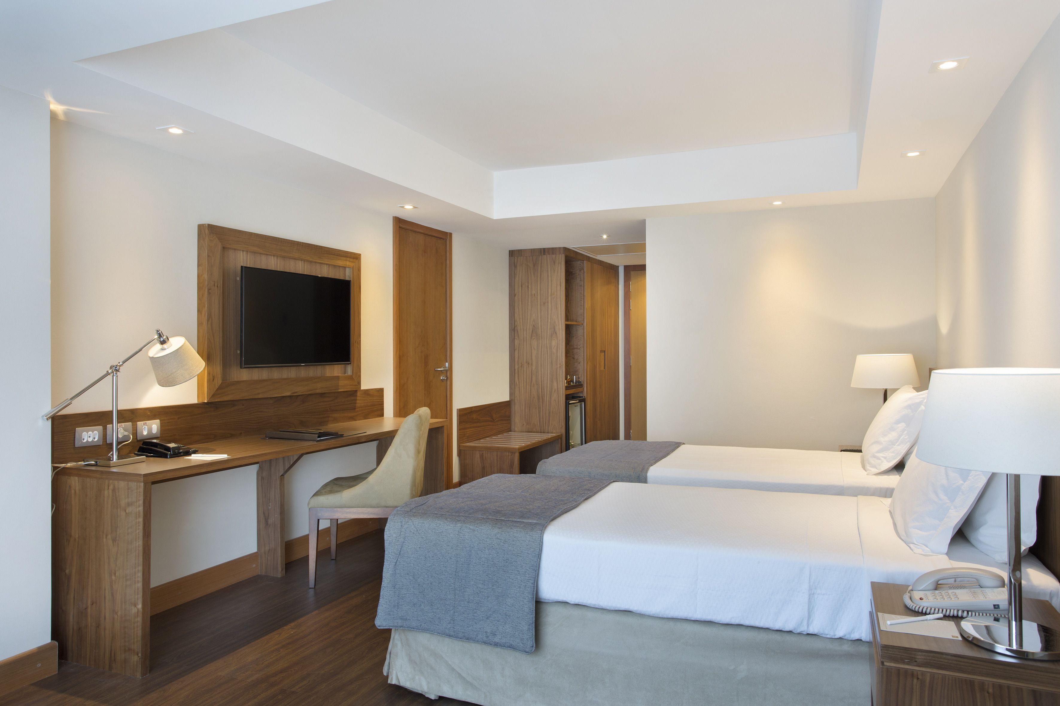 Et moderne hotelværelse med et træskrivebord, to senge, et fjernsyn og en skrivebordslampe.
