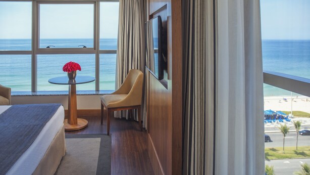 Una habitación de hotel con un ventanal grande, una cama, una silla, una mesita y un televisor.