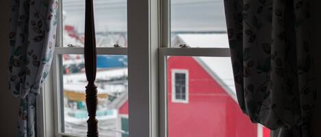 A room with a four-poster bed, a window with curtains, and a view of a red building outside.