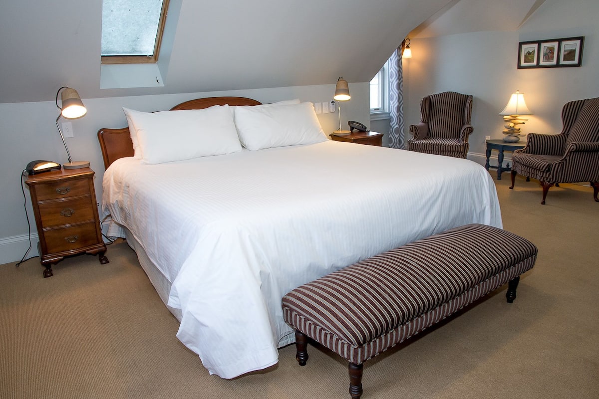 A neatly arranged bedroom with a large bed, bedside lamps, a wooden nightstand, a bench, and a chair.