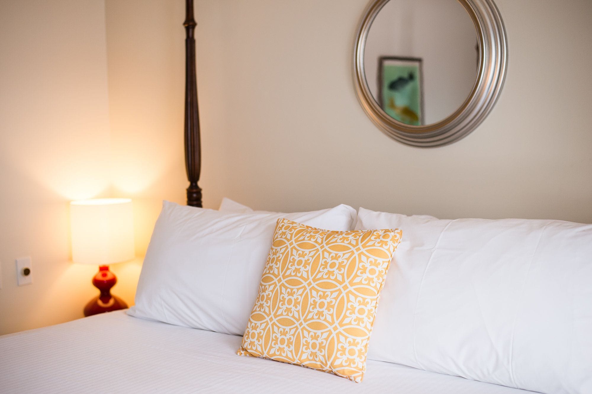 Cama bem arrumada com almofadas brancas e estampadas, lumin ária de cabeceira e espelho redondo na parede.