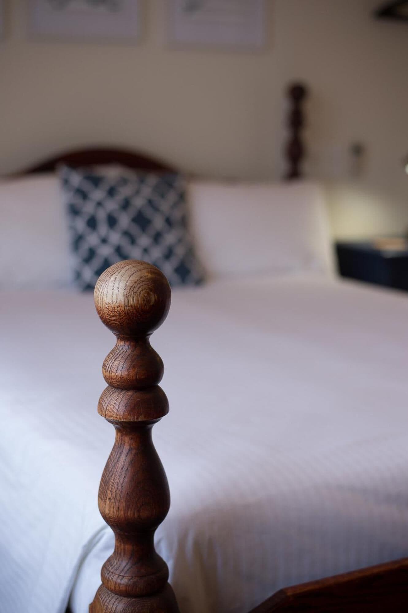 A wooden bedpost with a rounded top, a bed with white bedding, and a patterned pillow in the background.