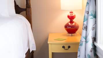 A neatly made bed with white linens, a wooden nightstand with a red lamp, and a patterned curtain.