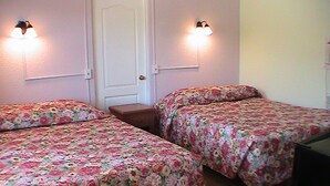 Huoneessa on kaksi sänkyä, joissa on molemmissa kukkakuviolliset vuodevaatteet, puinen yöpöytä ja seinään kiinnitetyt valaisimet.