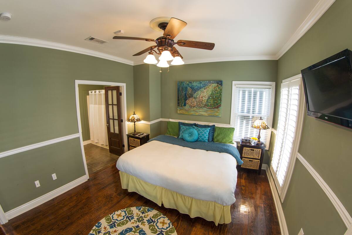 A bedroom with a large bed, a ceiling fan, a painting on the wall, and a TV mounted on the wall.