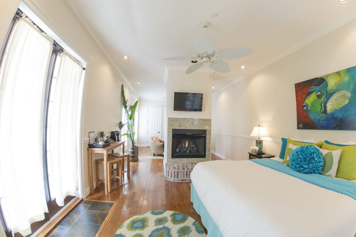 A bedroom with a bed, a fireplace, a ceiling fan, and a painting on the wall.
