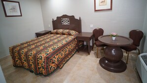 A bedroom with a bed, a bedside table, a chair, a round table, and a potted plant. - The Fly Inn (Lynwood)
