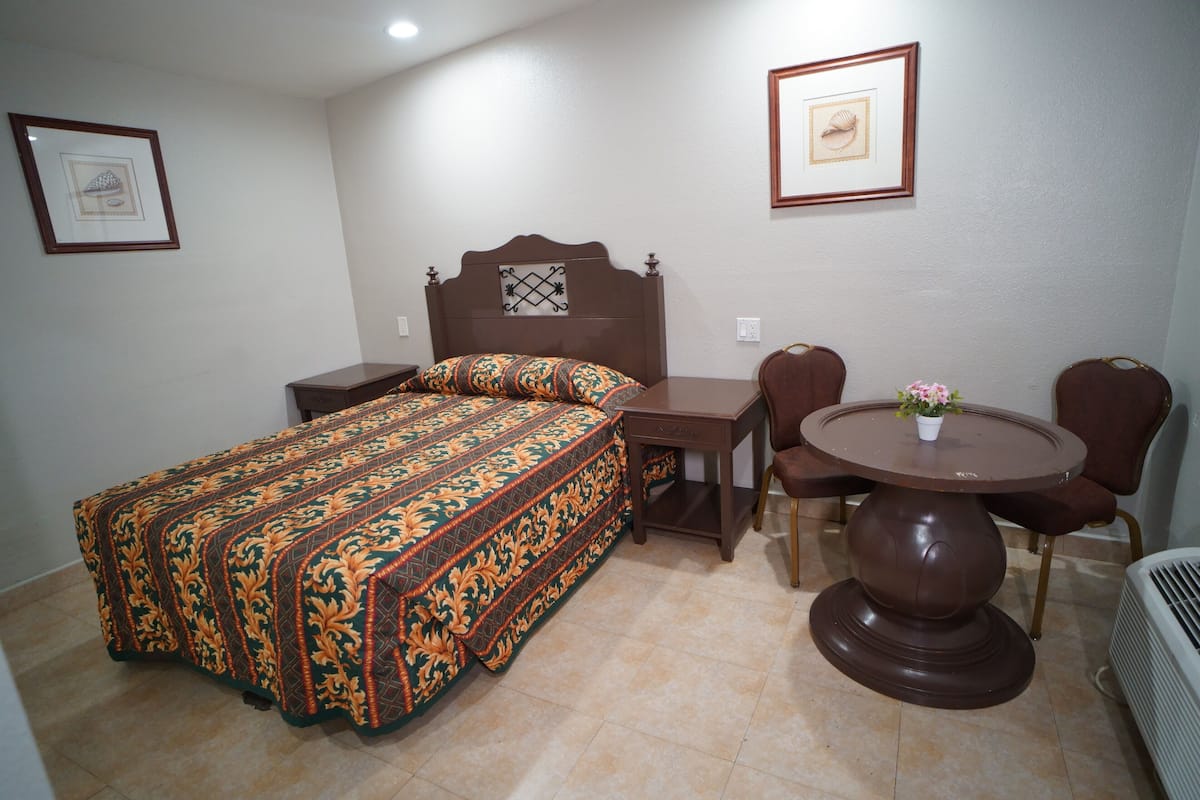 A bedroom with a bed, a bedside table, a chair, a round table, and a potted plant.