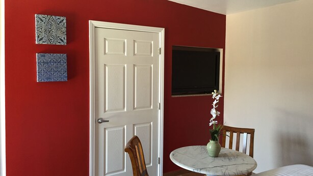 A room with a red wall, a white door, a small table with a vase of flowers, a chair, and a bed. - The Fly Inn (Lynwood)