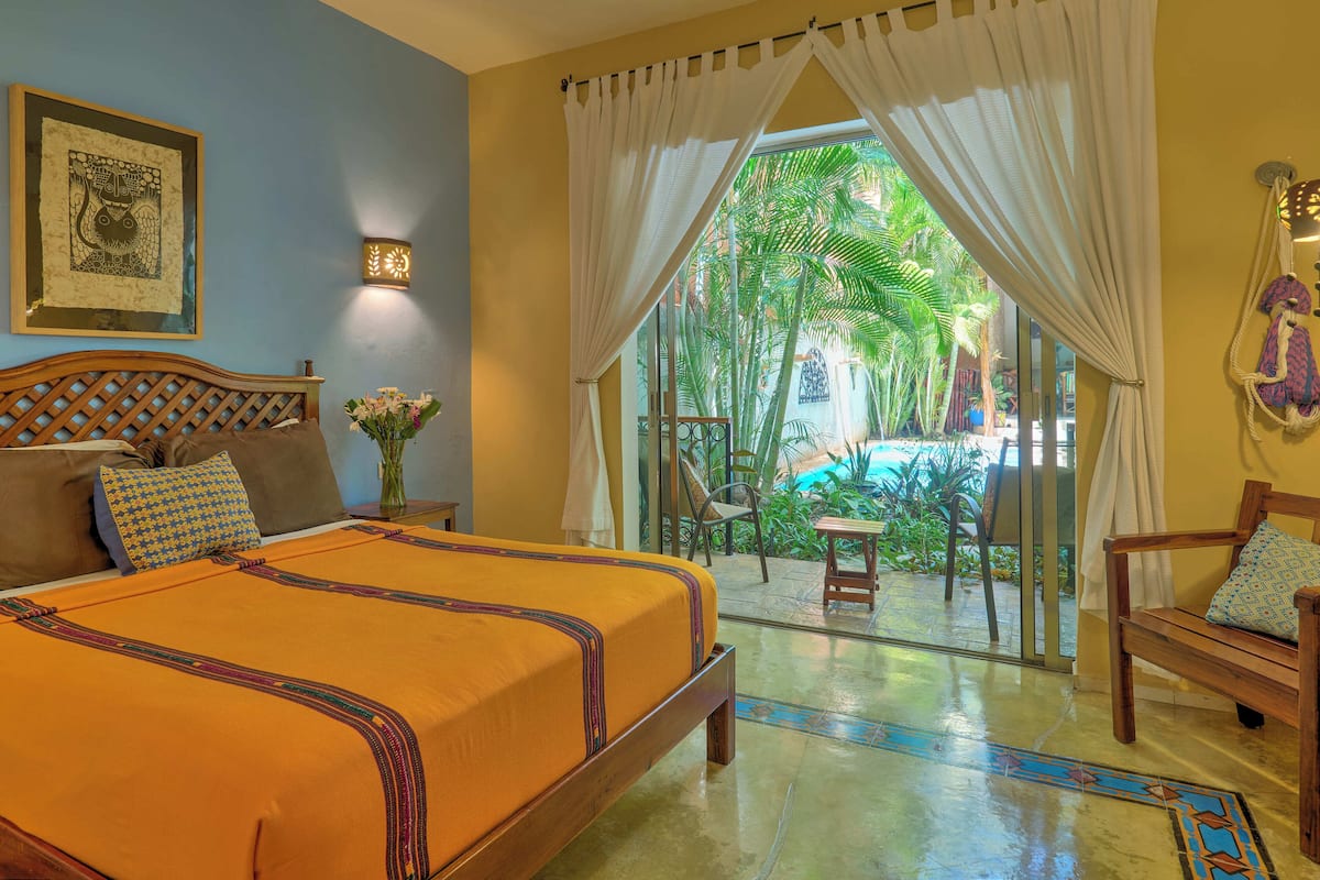 A hotel room with a bed, a nightstand, a vase with flowers, a framed picture on the wall, and a view of a pool and garden through the curtains.