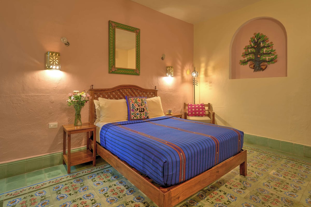 A bedroom with a wooden bed, floral-patterned floor tiles, a nightstand with a vase of flowers, and wall-mounted lights.