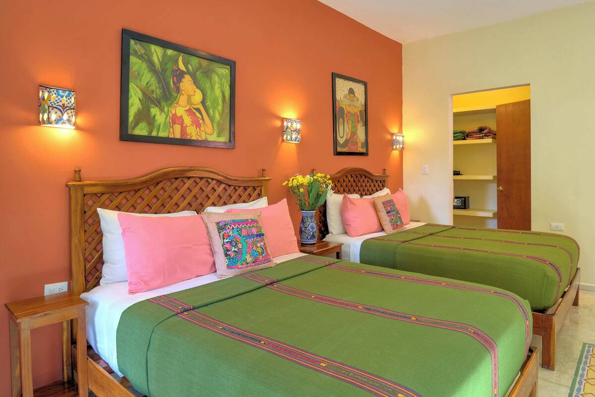 A hotel room with two beds, each with green bedspreads and pink pillows. There are wall-mounted lights and a painting above the beds.