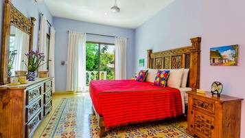 A bedroom with a red bedspread, wooden dresser, and a view of greenery outside.