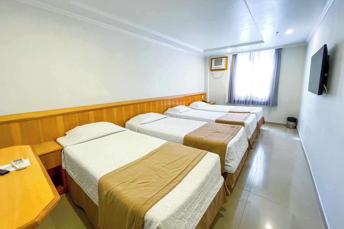 A hotel room with three single beds, a wooden headboard, a bedside table with a phone and a remote control, a TV mounted on the wall, and a window with curtains.