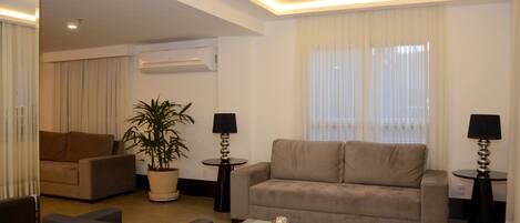 A modern living room with a beige sofa, a coffee table, and a potted plant. There are two lamps on side tables and a ceiling light fixture.