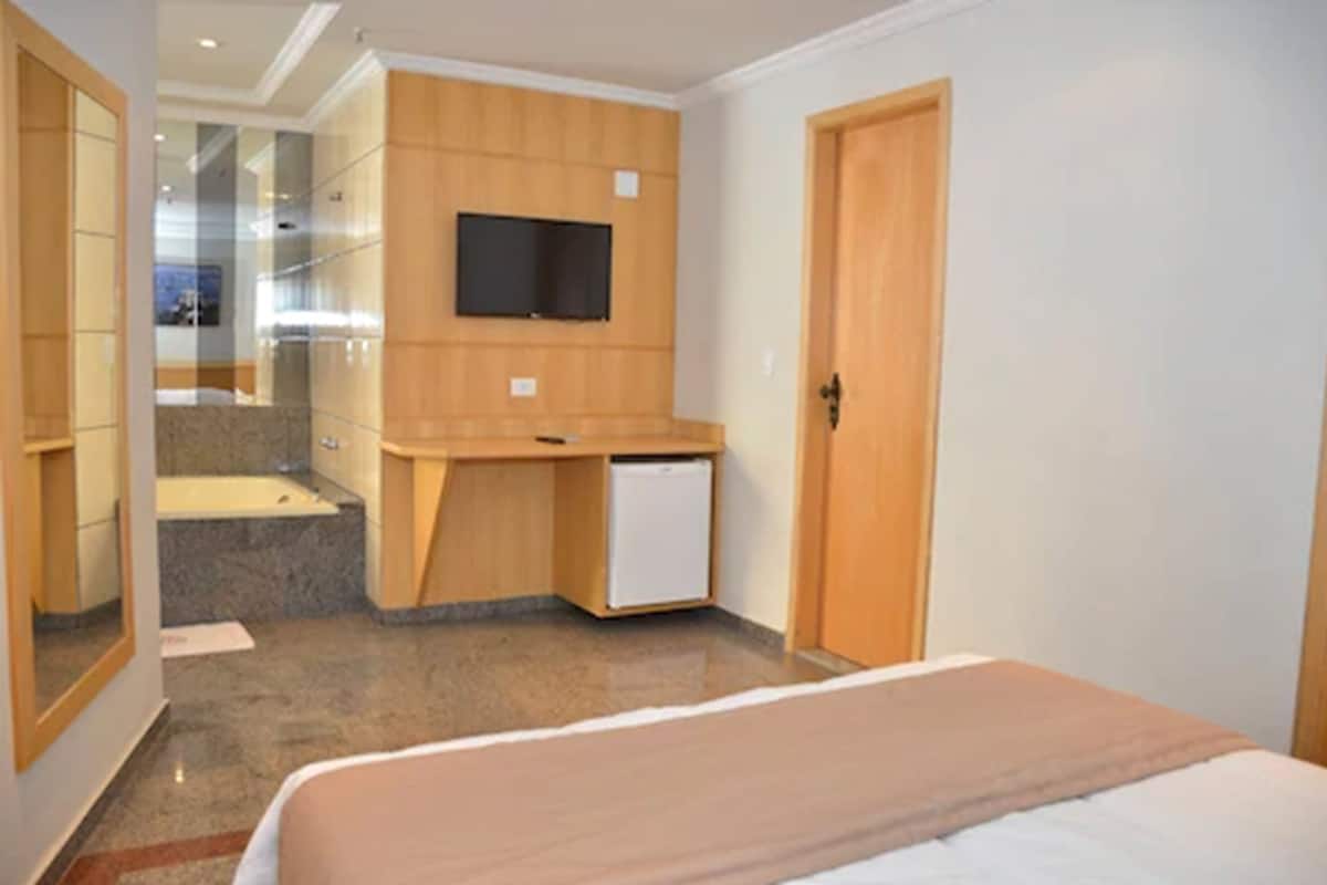 A hotel room with a bed, a small desk with a television, a mirror, and a door.