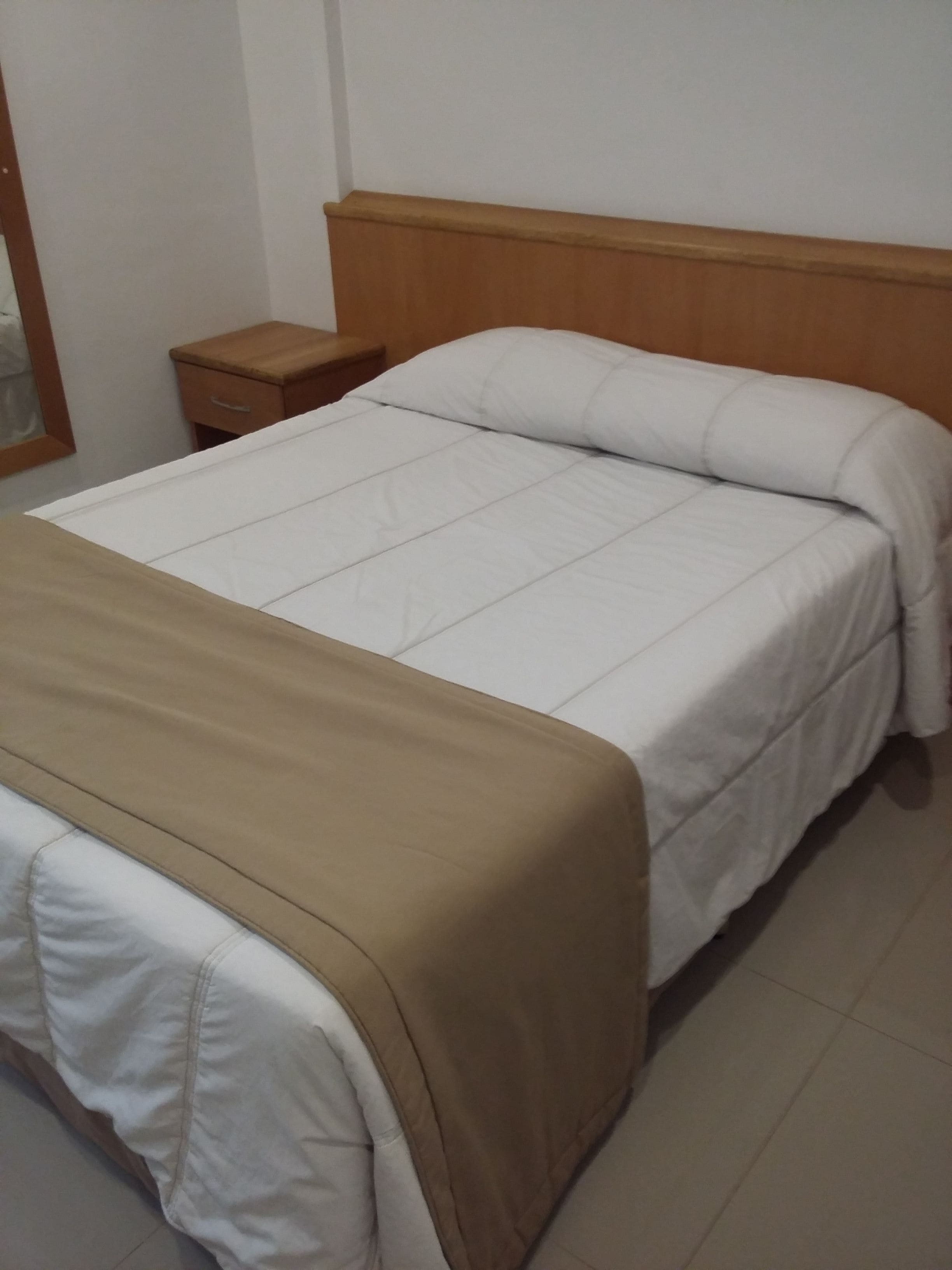 Uma cama com cabeceira de madeira, mesa de cabeceira e manta bege.