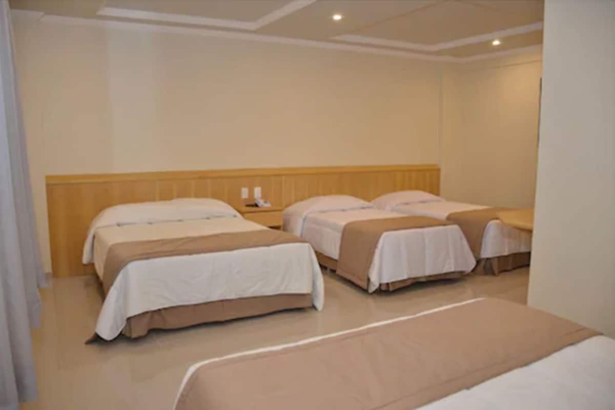 A hotel room with three beds arranged in a row, a wooden headboard, and a small table with a lamp.