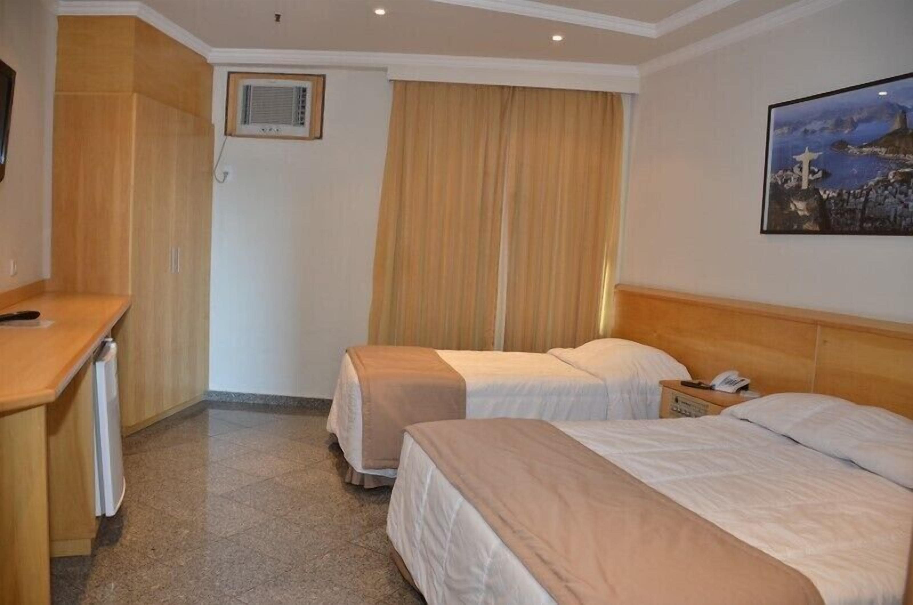A hotel room with two beds, a desk, a chair, a window with curtains, and a painting on the wall.