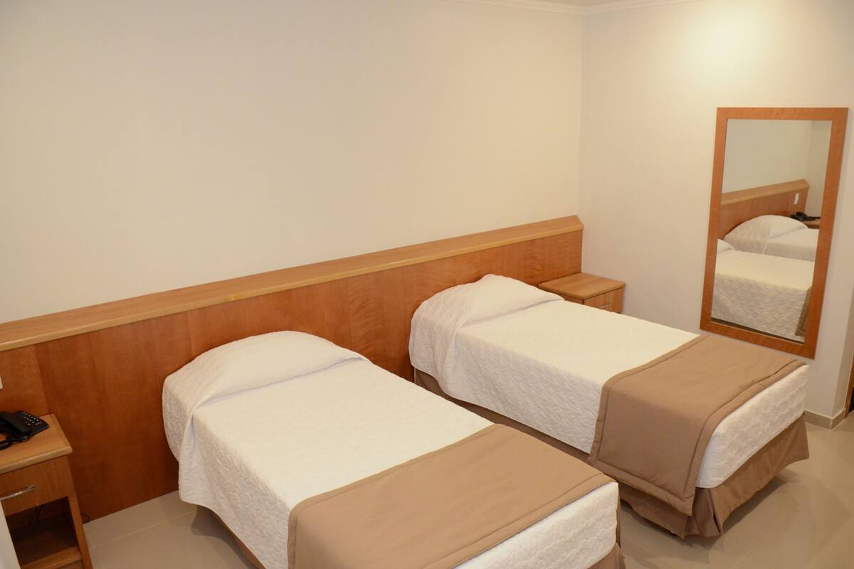 A hotel room with two single beds, a wooden headboard, a bedside table with a phone, and a mirror on the wall.
