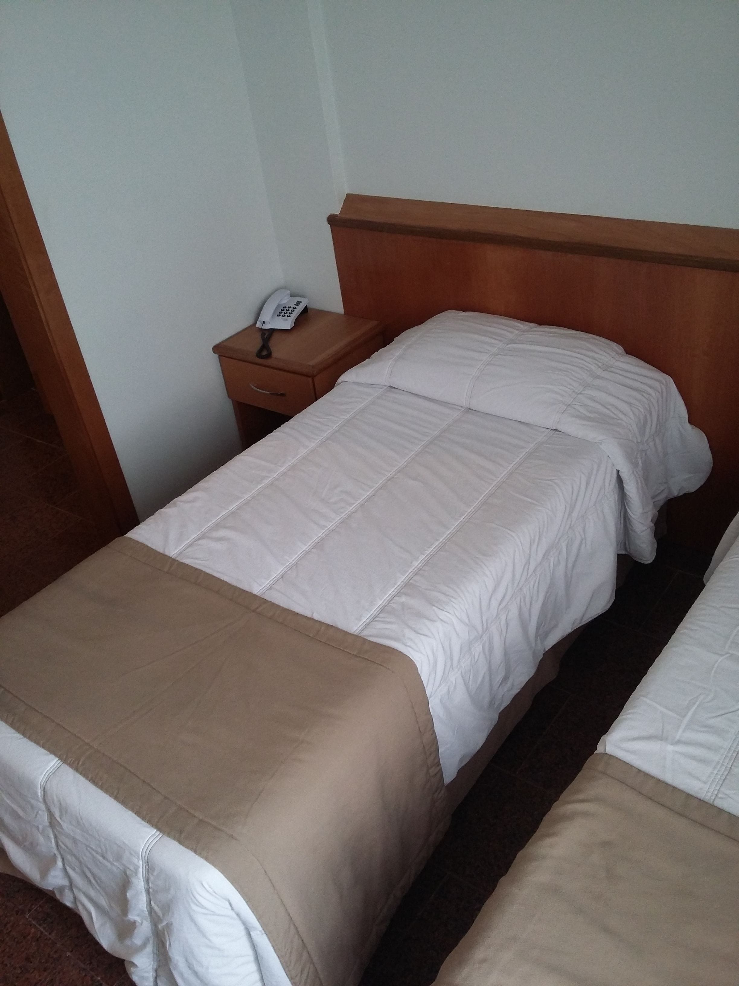 A hotel room with two single beds, a wooden headboard, a bedside table with a telephone, and a brown bedside table.