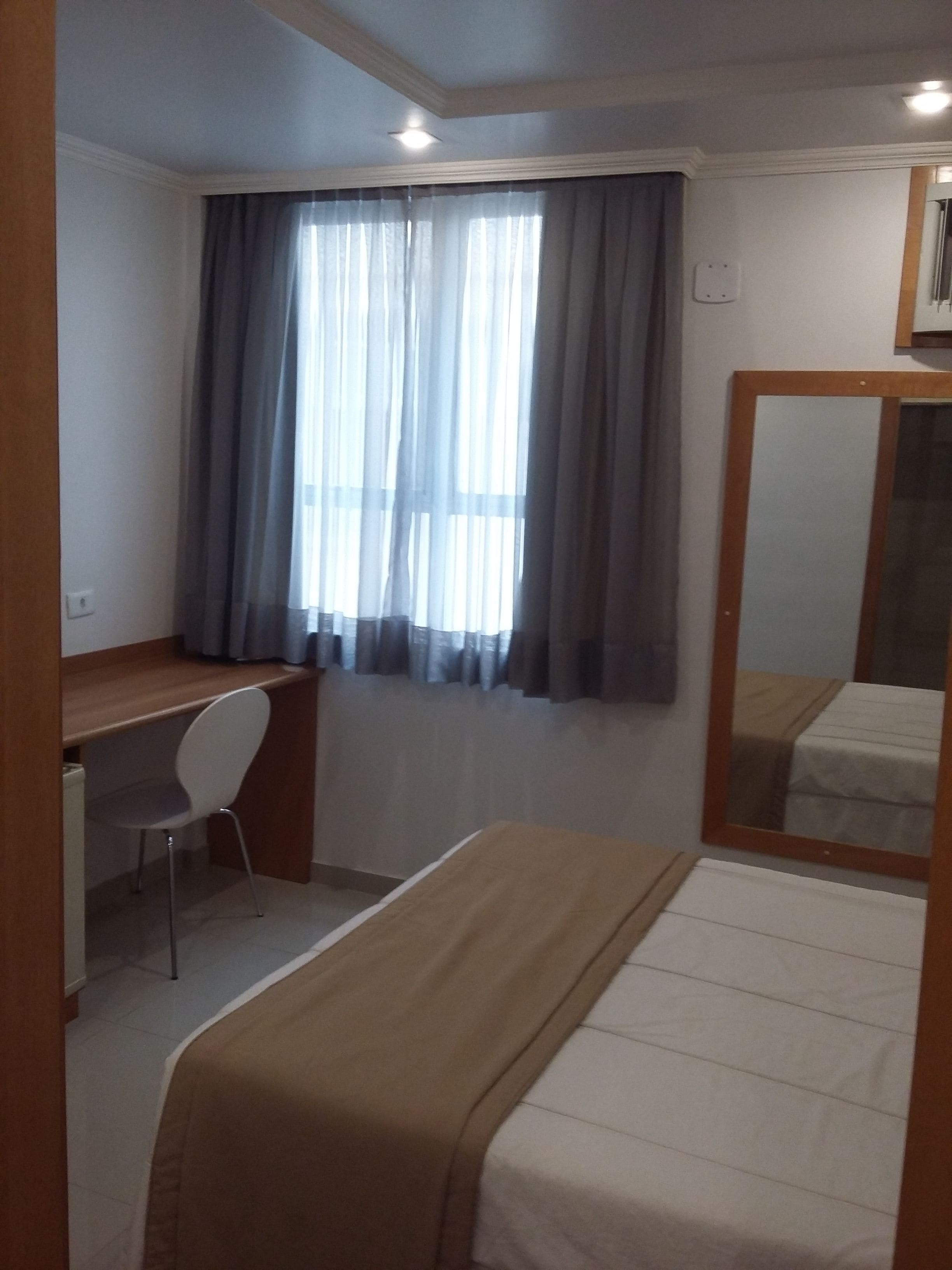 A small, well-lit room with a single bed, a desk, and a chair. There is a large mirror on the wall, and a window with curtains.