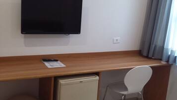 Una habitación pequeña con un escritorio de madera, un televisor fijado a la pared, un refrigerador pequeño, una silla blanca y una ventana con cortinas.