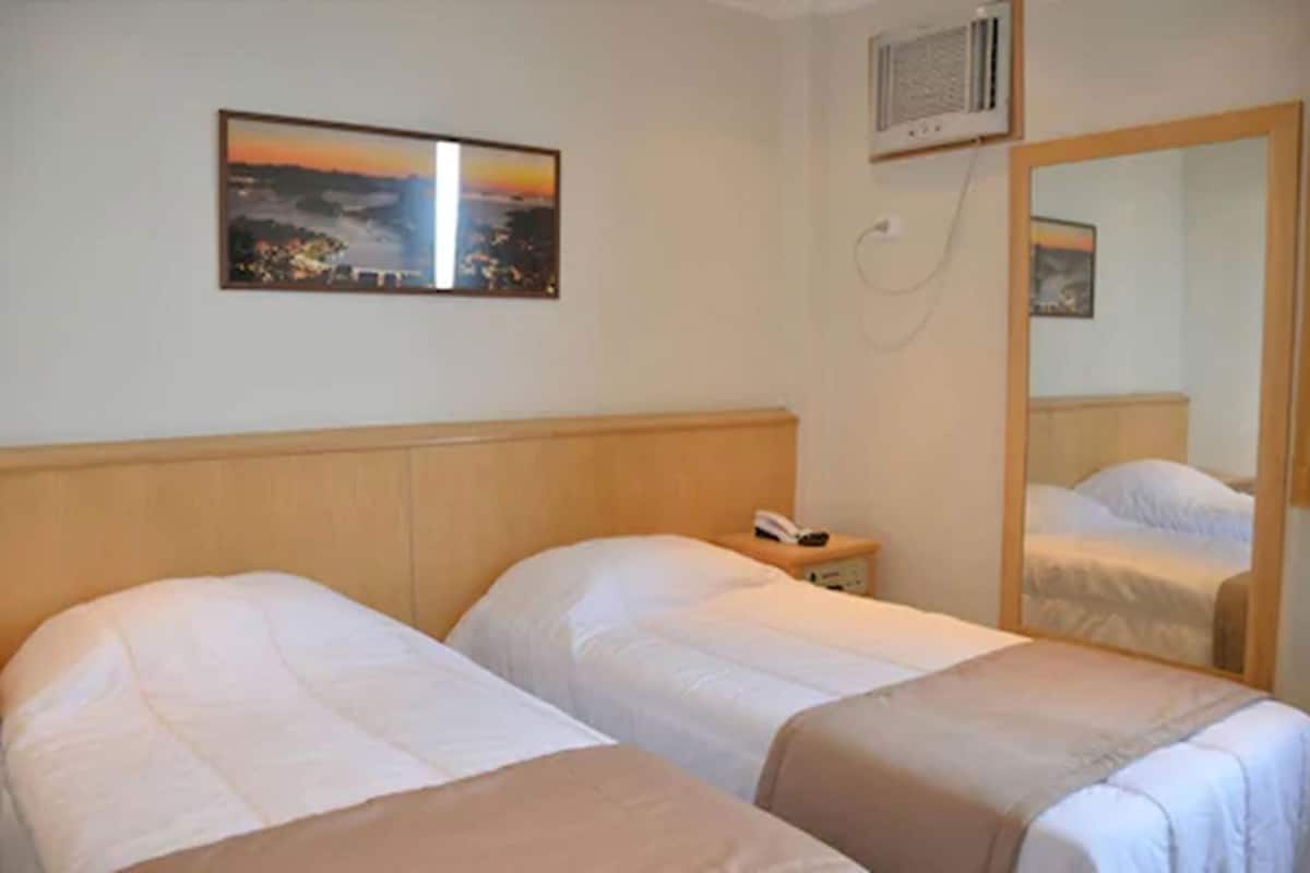 A hotel room with two beds, a nightstand with a phone, a wall-mounted air conditioner, and a framed picture above the beds.