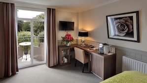 Une chambre d’hôtel équipée d’un bureau, d’une chaise, d’une petite table avec une lampe, d’un radiateur, d’une télévision et d’une grande fenêtre avec des rideaux.