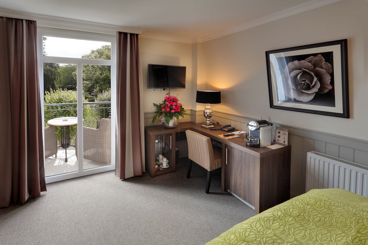 A hotel room with a desk, chair, a small table with a lamp, a radiator, a TV, and a large window with curtains.