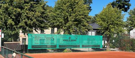 Una cancha de tenis con una cerca y un cartel anunciando Ribs & Bones Steakhouse, Wings & Drums y Belon.