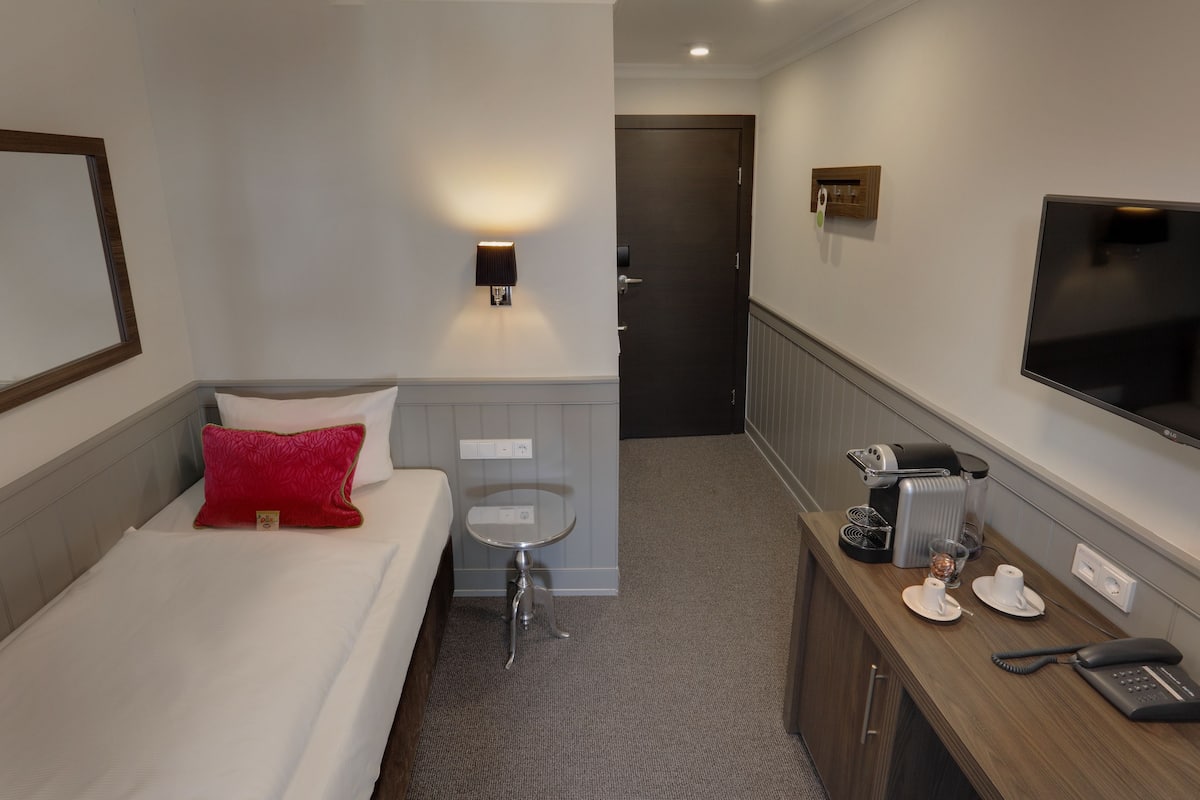 A hotel room with a bed, a small table with a lamp, a TV mounted on the wall, and a coffee maker.