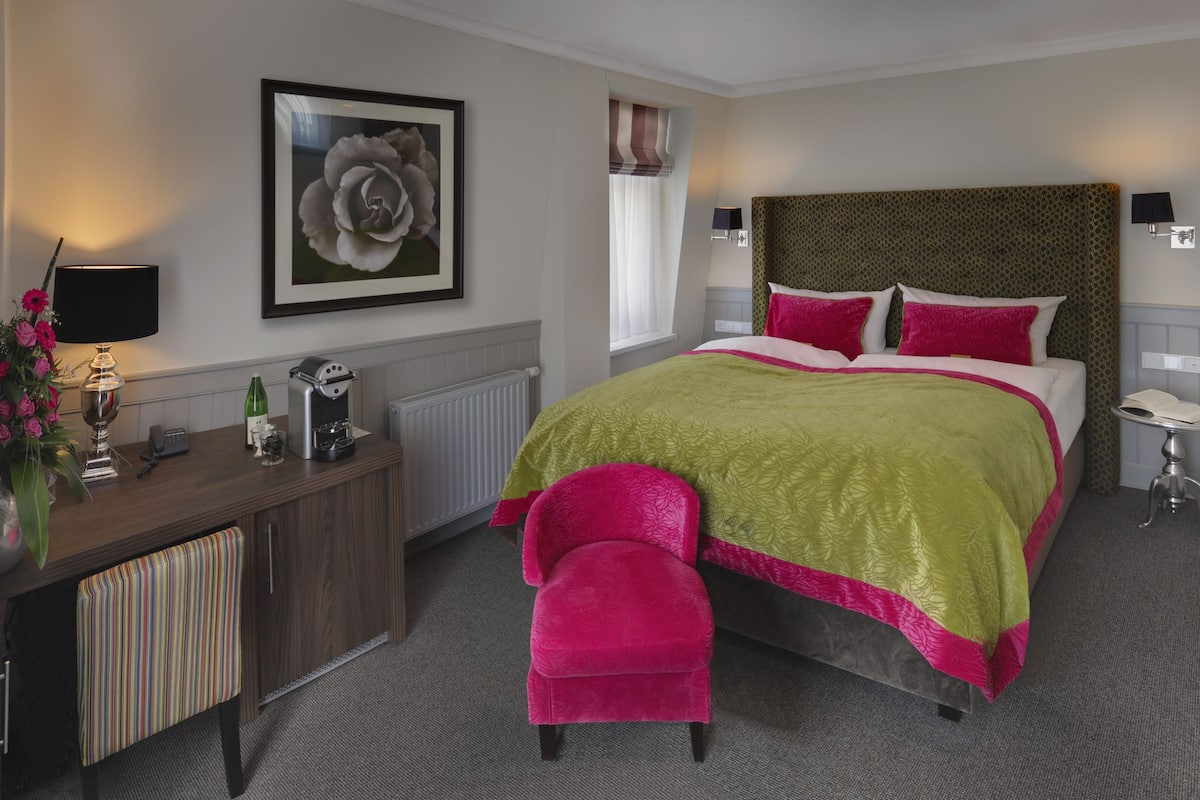 A hotel room with a large bed, a desk with a lamp, a chair, and a framed picture on the wall.