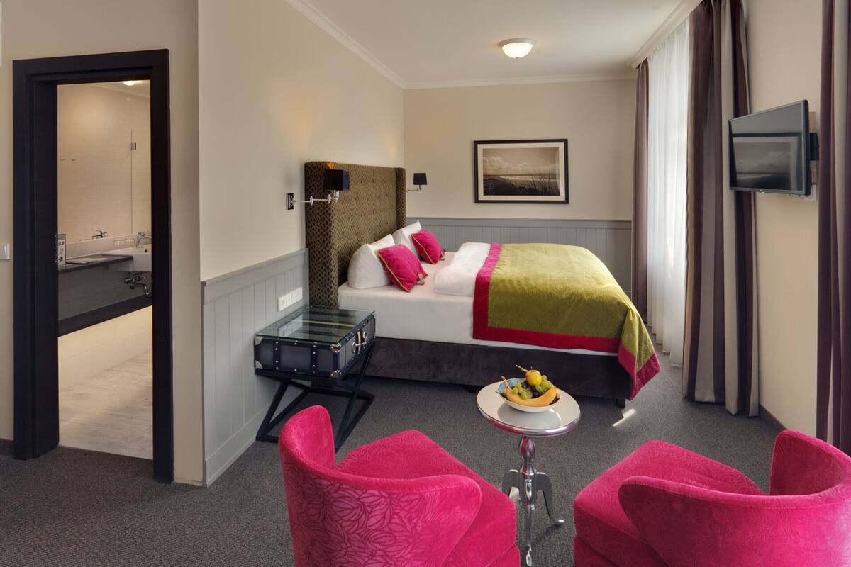 A hotel room with a bed, a small table with a fruit bowl, and two pink chairs.