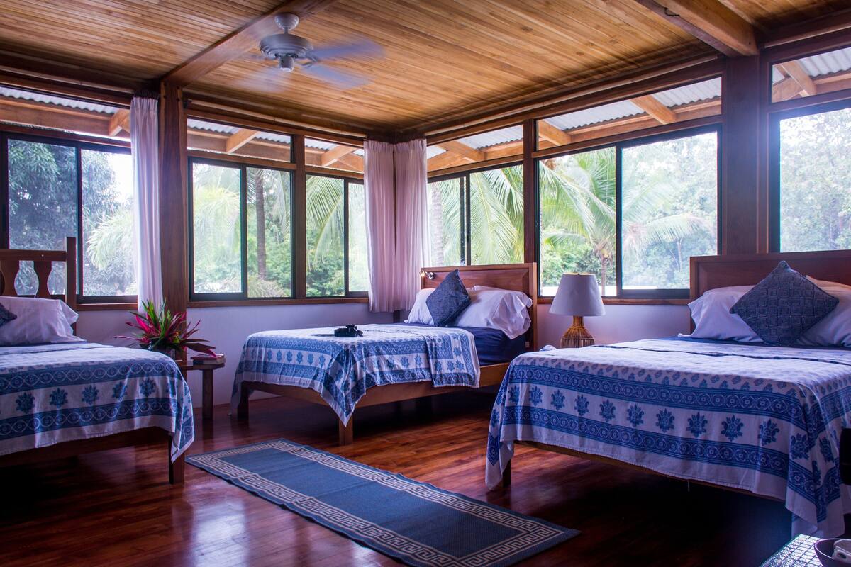 Habitación con tres camas, piso de madera, ventanas amplias y ventilador de techo.