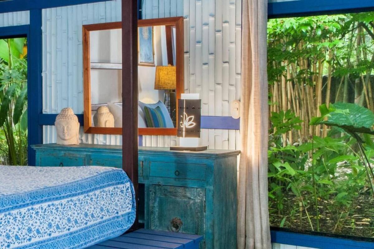Un dormitorio con cama, mesita de noche, lámpara y espejo. Se ve follaje verde a través de la ventana.