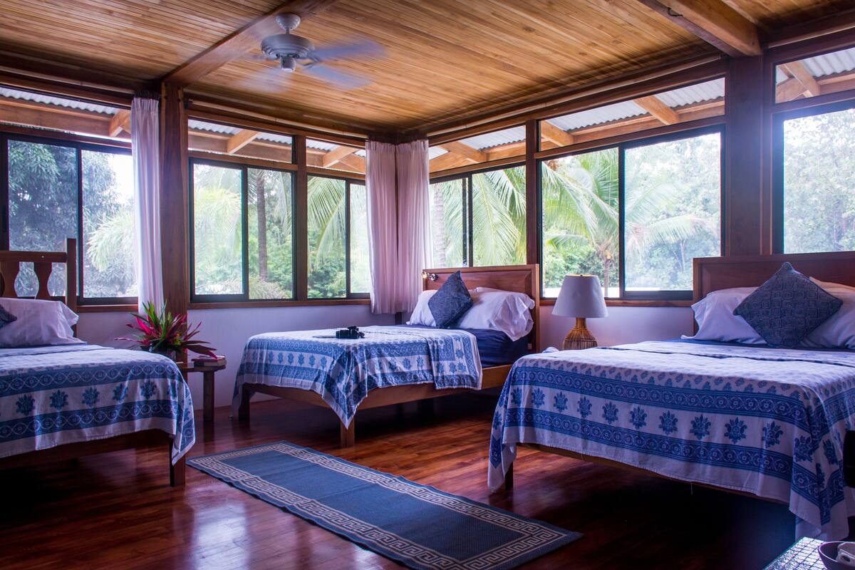 Una habitación con tres camas, piso de madera, ventanas amplias y un ventilador de techo.