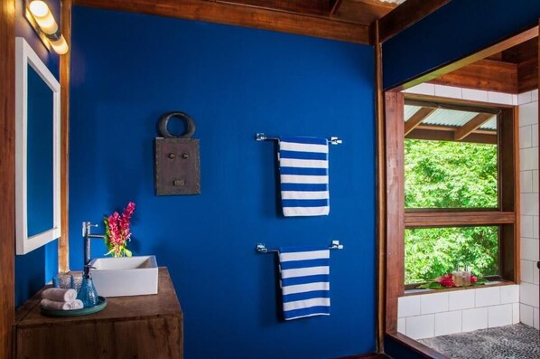 A bathroom with a blue wall, wooden ceiling, and a window with a view of greenery. There are two striped towels hanging on hooks and a white sink with decorative items.
