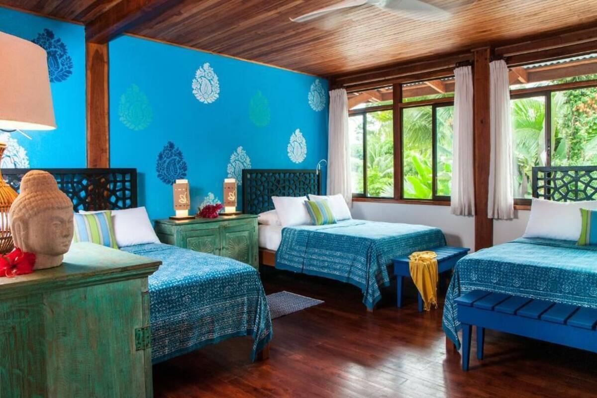 Una habitación con cuatro camas, cada una con una colcha azul estampada, cabeceras de madera y un gran ventanal con cortinas.