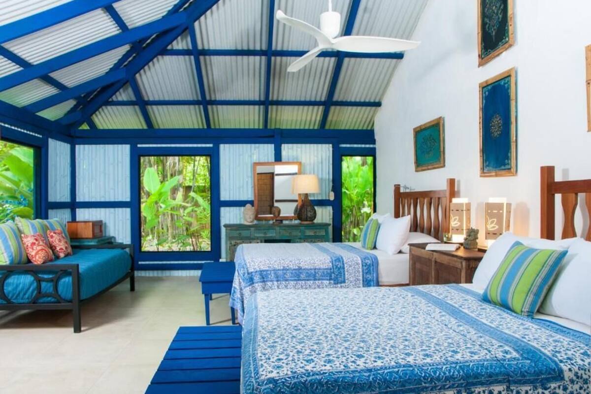 Una habitación con techos altos, una cama con ropa de cama azul, un banco de madera, un escritorio con una lámpara y vistas a zonas verdes al exterior.