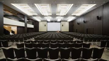 Une grande salle de conférence moderne, avec des rangées de chaises noires, un grand écran à l’avant et un escalier sur le côté gauche.