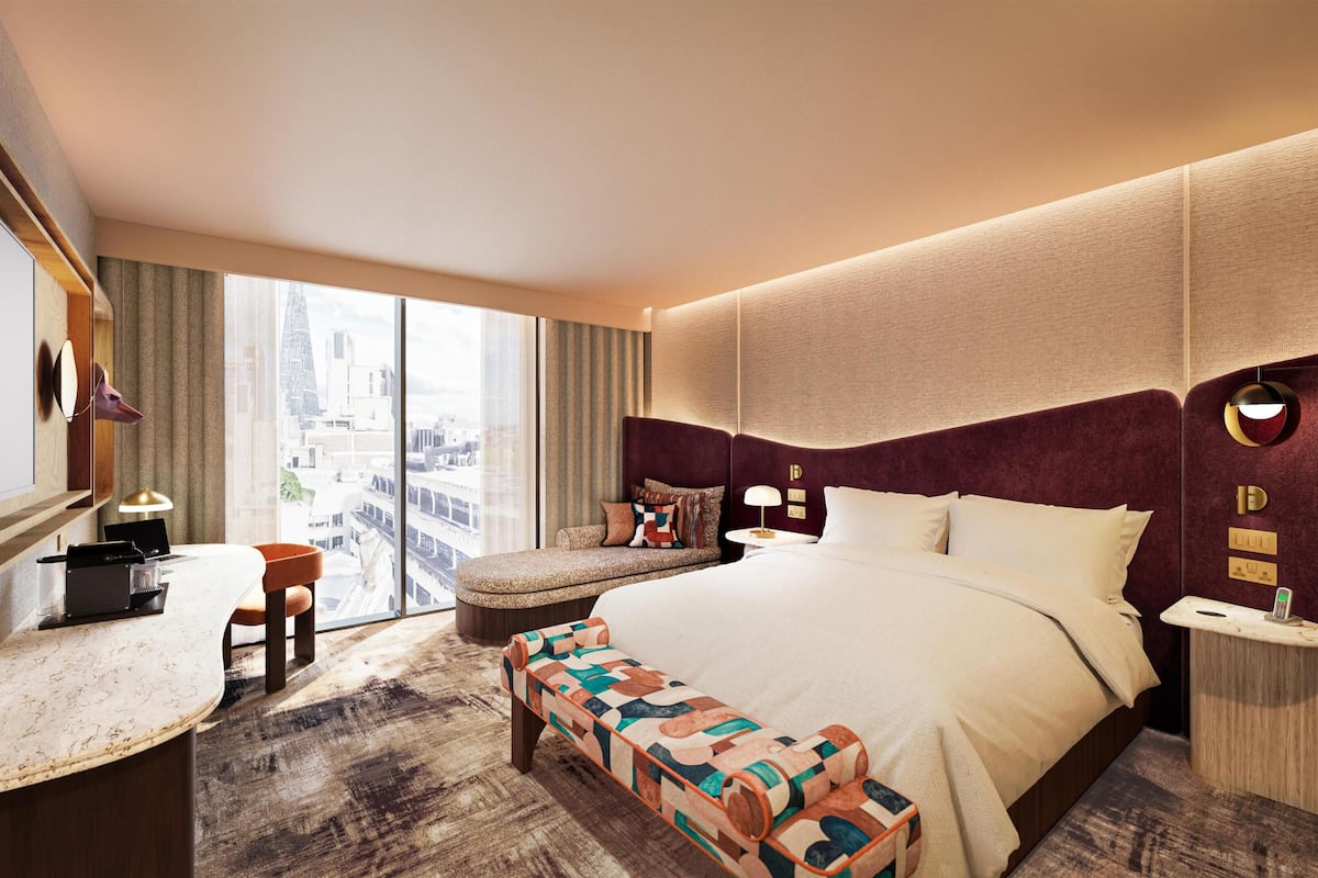 Habitación de hotel con cama, escritorio, silla, banco, mesitas de noche y vistas a la ciudad.