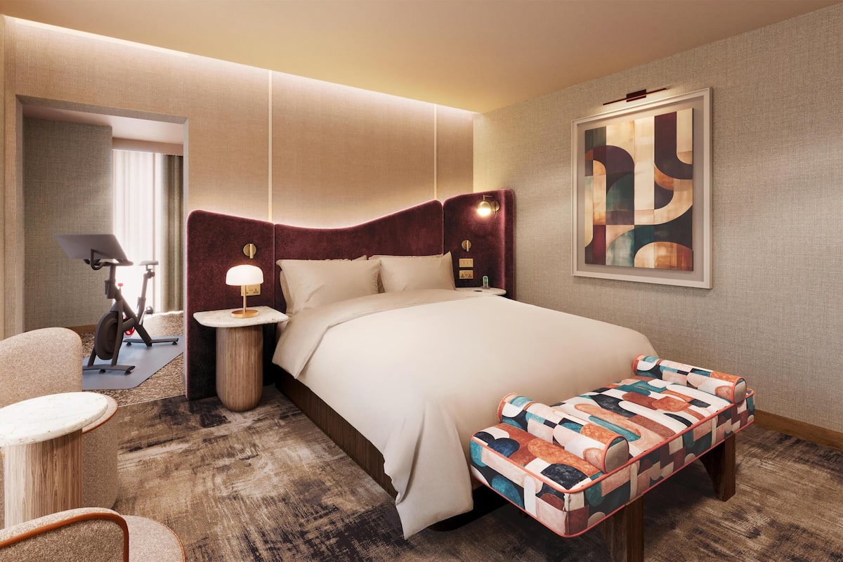 Habitación de hotel con una cama grande, un escritorio con lámpara, una silla, una cinta de correr y un banco colorido.