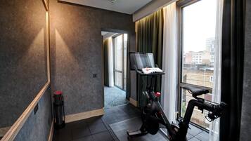 A modern hotel room with a large window, a ceiling-mounted fire alarm, a wall-mounted television, and a Peloton exercise bike.