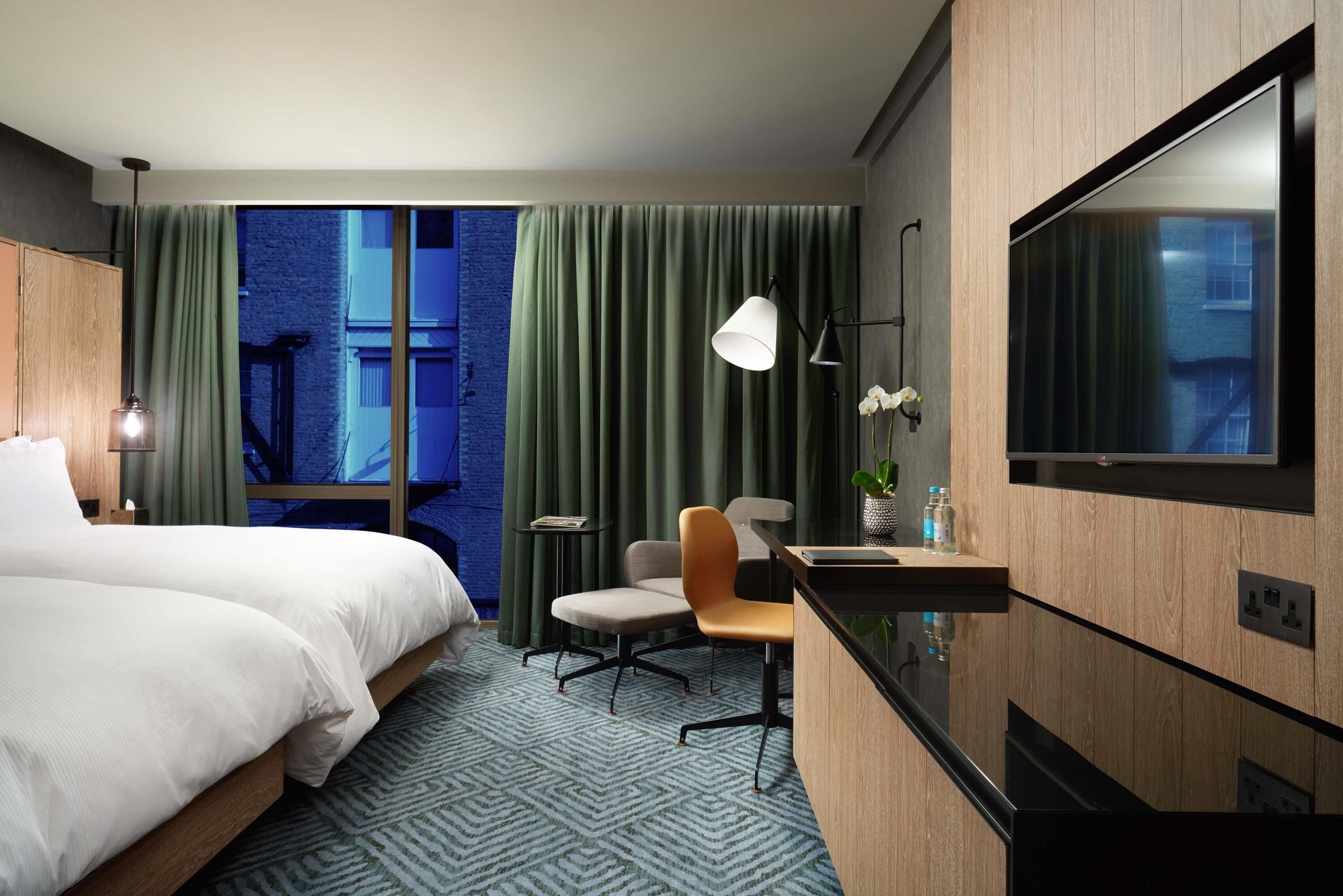A hotel room with a large bed, a desk with a chair, a TV, and a view of a building through the window.