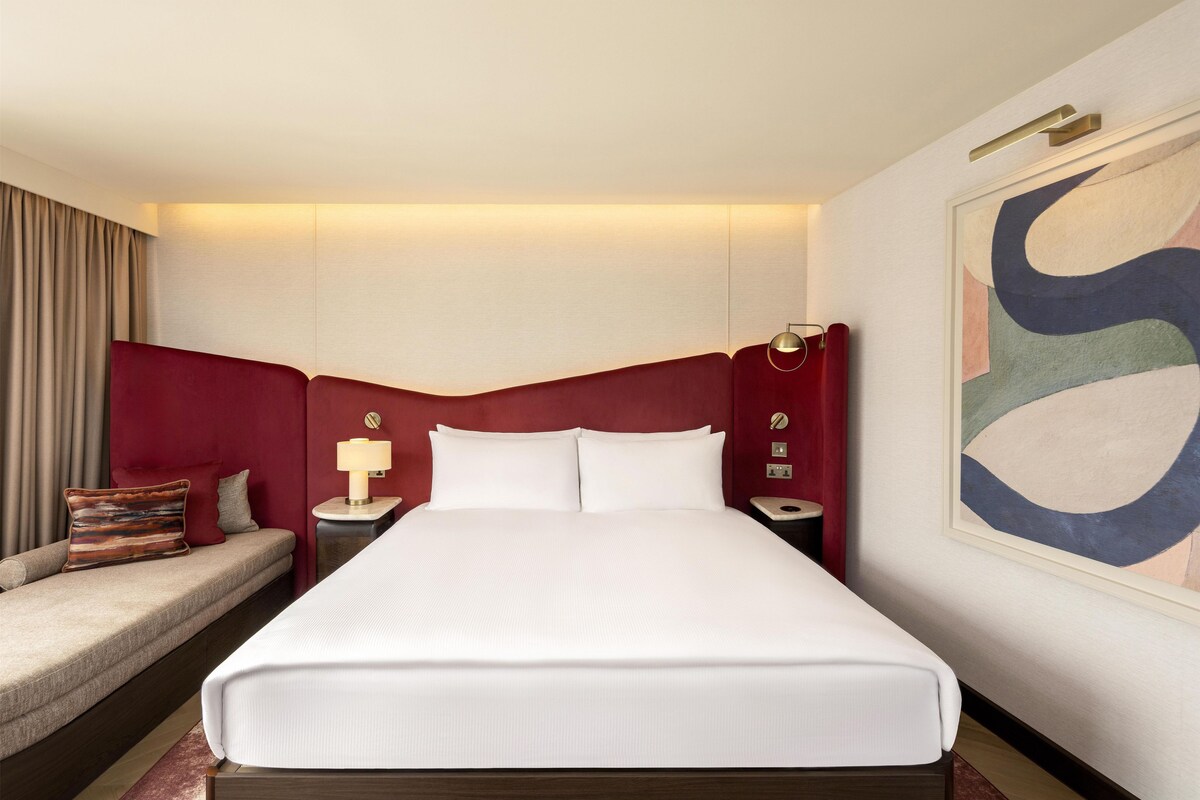 Habitación de hotel con una cama grande, una mesita de noche con una lámpara, cabecero rojo, una obra de arte en la pared y un sofá con un cojín.