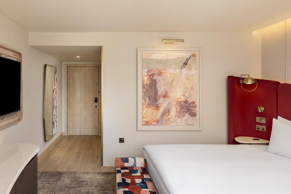 Habitación de hotel con cama, televisor, un cuadro y cabecero rojo.