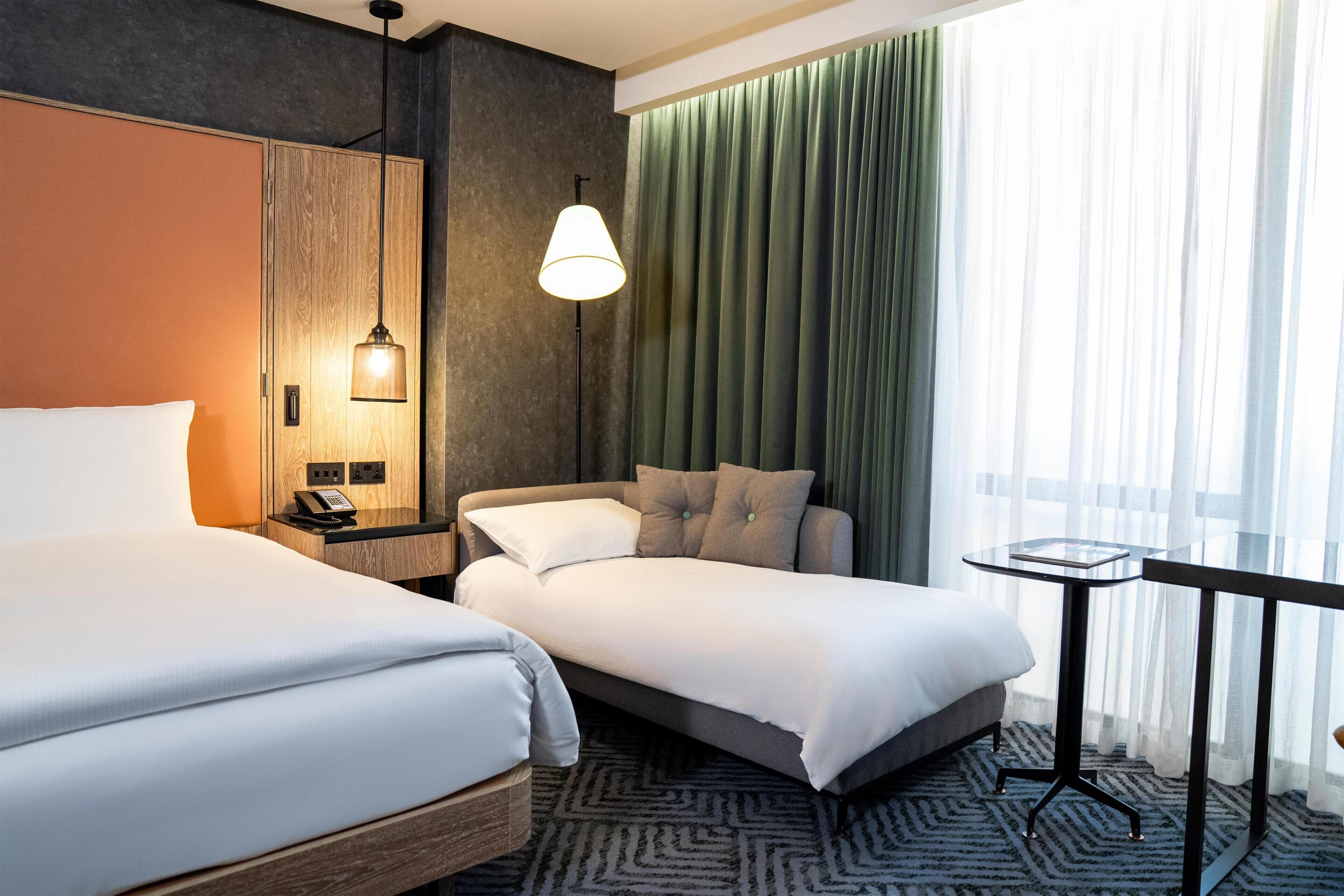 Habitación de hotel con dos camas, una mesita de noche con lámpara, una pequeña mesa y vistas al exterior a través de cortinas translúcidas.