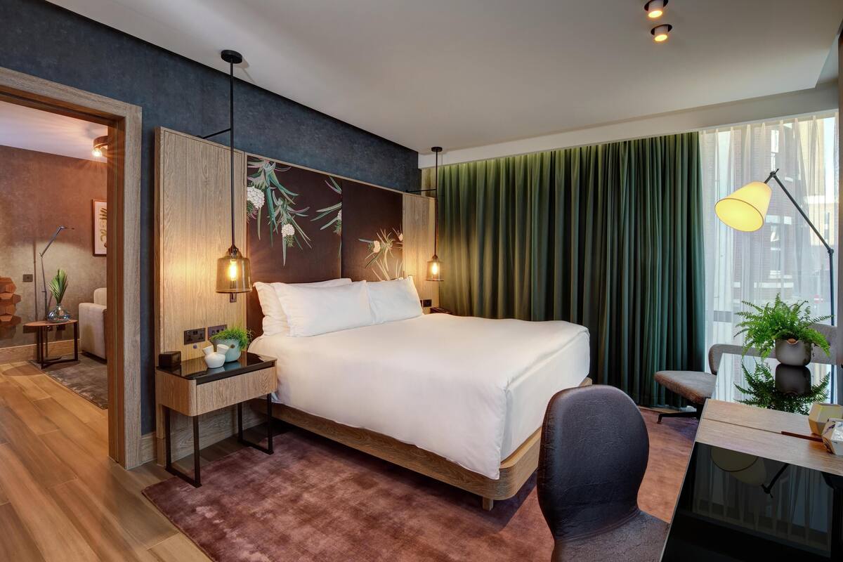 A modern hotel room with a bed, bedside table, lamp, and a view of the city through large windows.