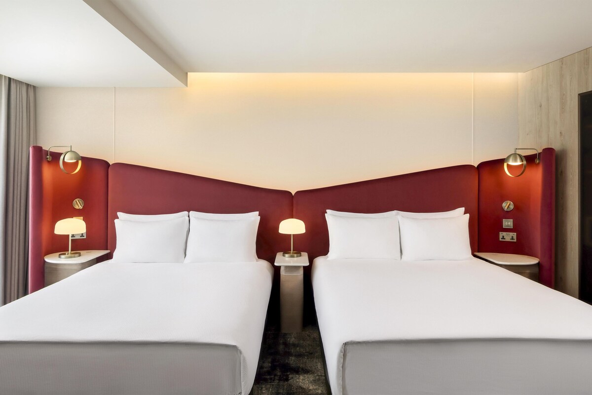 Habitación de hotel con dos camas, una mesita de noche con lámpara y un espejo grande.